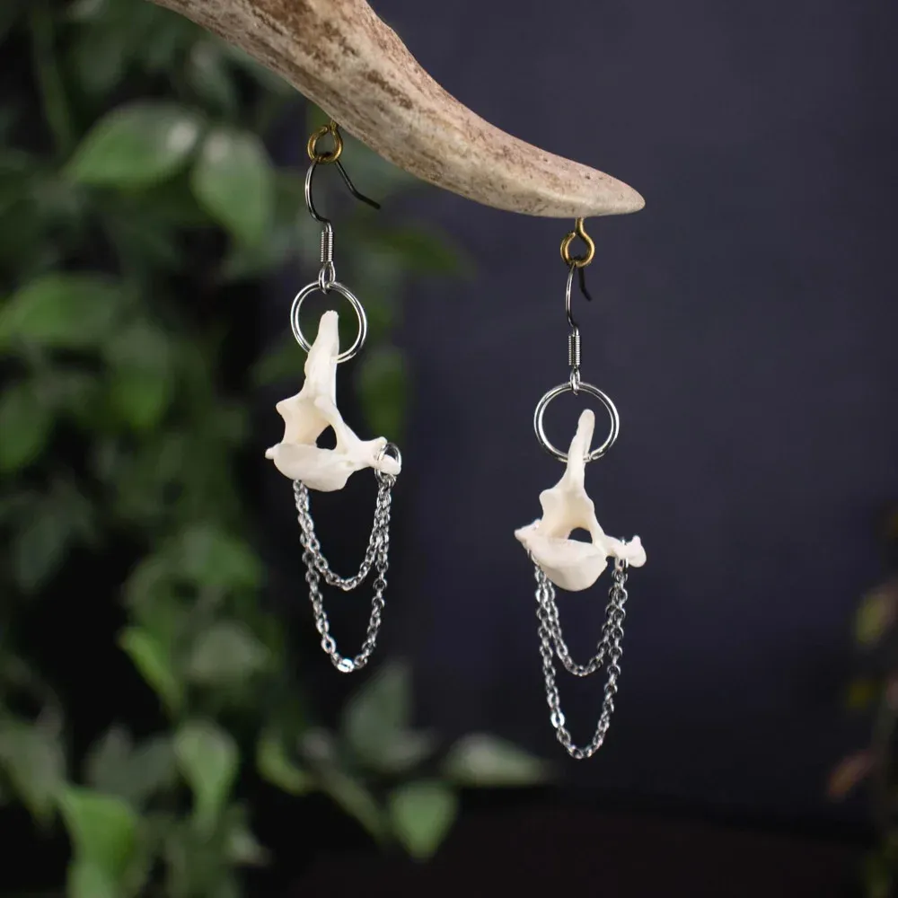 Gothic style dangled earrings with white marten bone vertebrae and thin silver chains hanging elegantly against a shadowy forest backdrop.