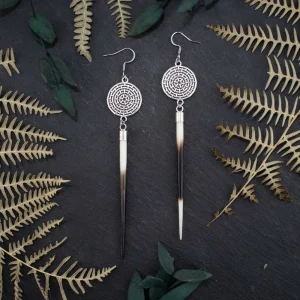 Detailed shot of silver quill earrings featuring round metallic charms and real porcupine quills, styled on a dark textured slate background framed by shadowy dried ferns.