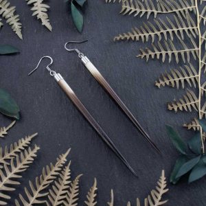 Alternative style long quill earrings resting on a dark stone slab, surrounded by the organic curves of dried ferns in a moody, low-light setting.