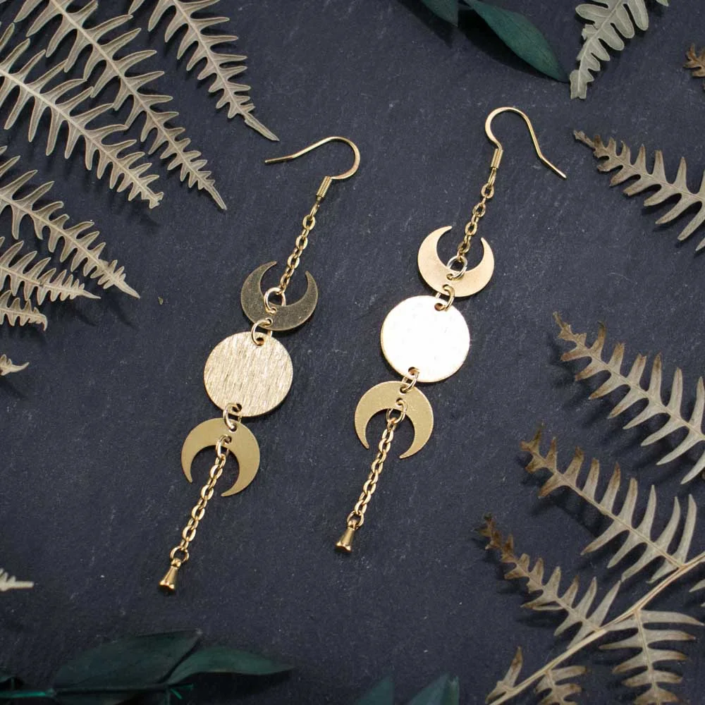 Close-up of gold-toned celestial hoops featuring the full lunar cycle and polished metallic finishes, displayed on a dark, textured surface.